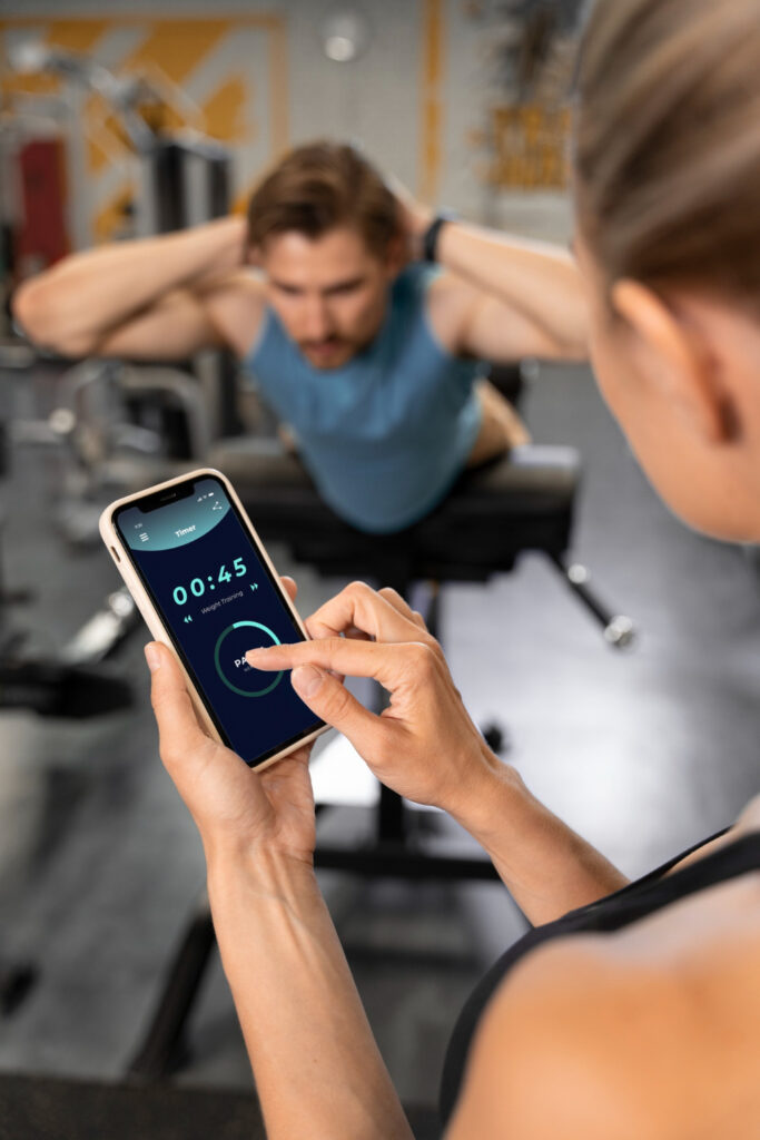 Triainwell fitness reviews: Woman holding smartphone with workout timer app while man performs back extensions at the gym.