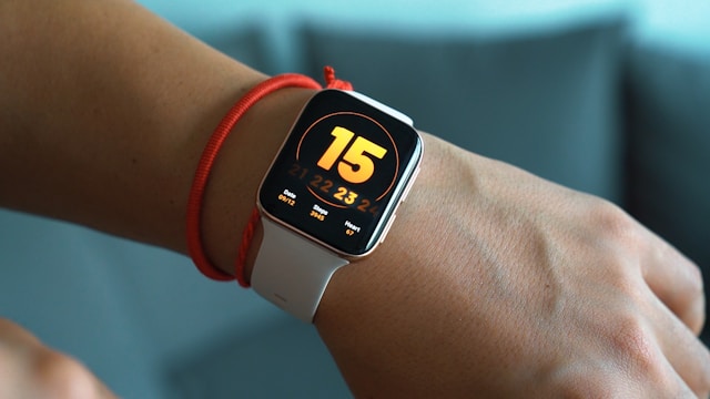 Future App Review: A white smartwatch with a bright 15 on the display and a red bracelet on the wrist, against a blue-gray blurred background.