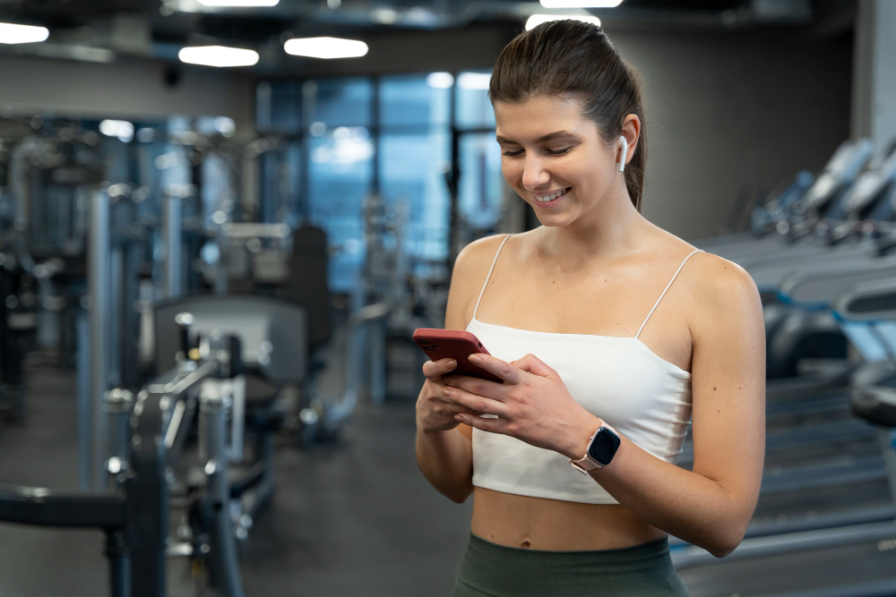 A young woman in a white crop top using a smartphone in a modern gym, with fitness equipment in the background.