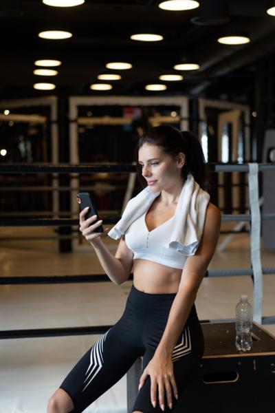 Female athlete sitting in boxing gym, checking smartphone after workout with towel over shoulders.