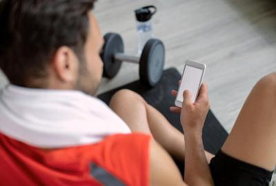 Male fitness coach sitting on mat checking smartphone after workout, dumbbell and bottle in background