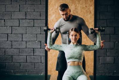 FitBudd Review: Personal trainer assisting female client with shoulder press exercise in gym.