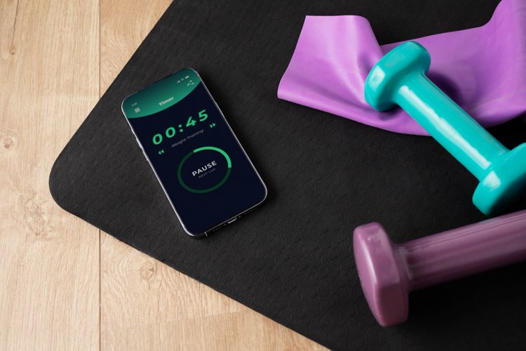 A smartphone on a black yoga mat displaying a workout timer app set for weight training, surrounded by colorful dumbbells and a purple resistance band on a wooden floor.