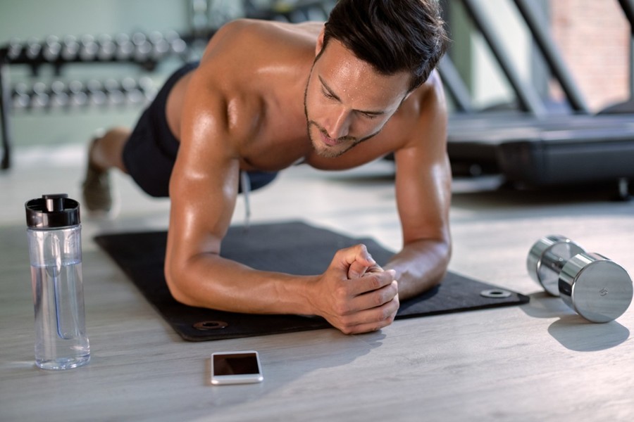 My PT Hub App: A fit man doing a plank on a mat in a modern gym, with a smartphone, dumbbell, and water bottle beside him.