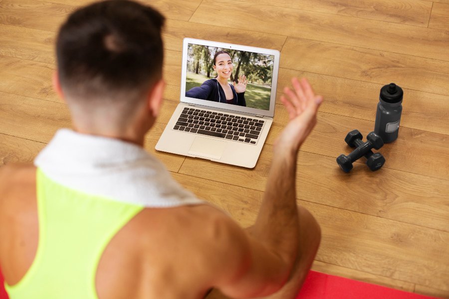 Man participating in online fitness coaching session via video call, with laptop, dumbbells, and water bottle on wooden floor.