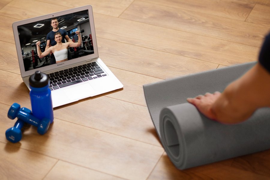 Trainerize Review: Laptop displaying online personal trainers during virtual workout session, with dumbbells, water bottle, and yoga mat on wooden floor.