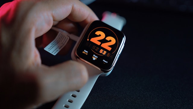 A person’s hand adjusting a white smartwatch with an orange digital display showing the number 22 and the current date and time.