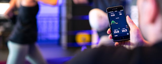 A person holding a smartphone with a fitness tracking app displayed, while others are exercising in a gym with blue lighting in the background.