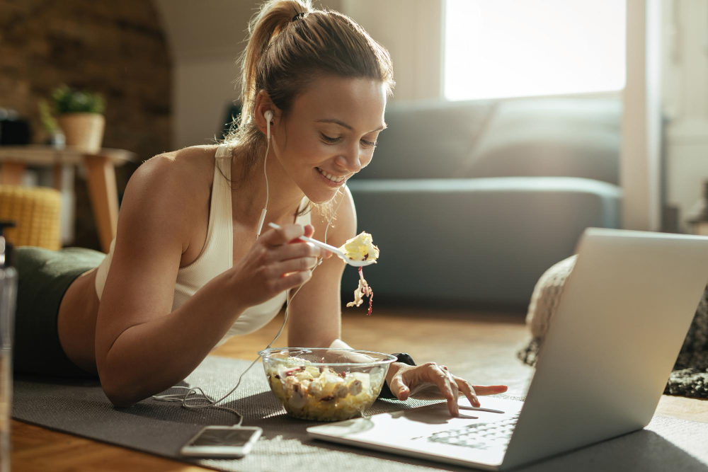 Active woman on yoga mat enjoying a nutritious lunch while streaming fitness training on her laptop.