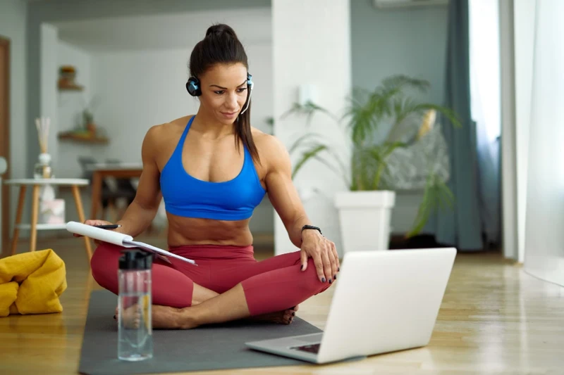 A fit online fitness coach sitting on a yoga mat, wearing a headset, coaching clients via laptop in a modern home gym setting.