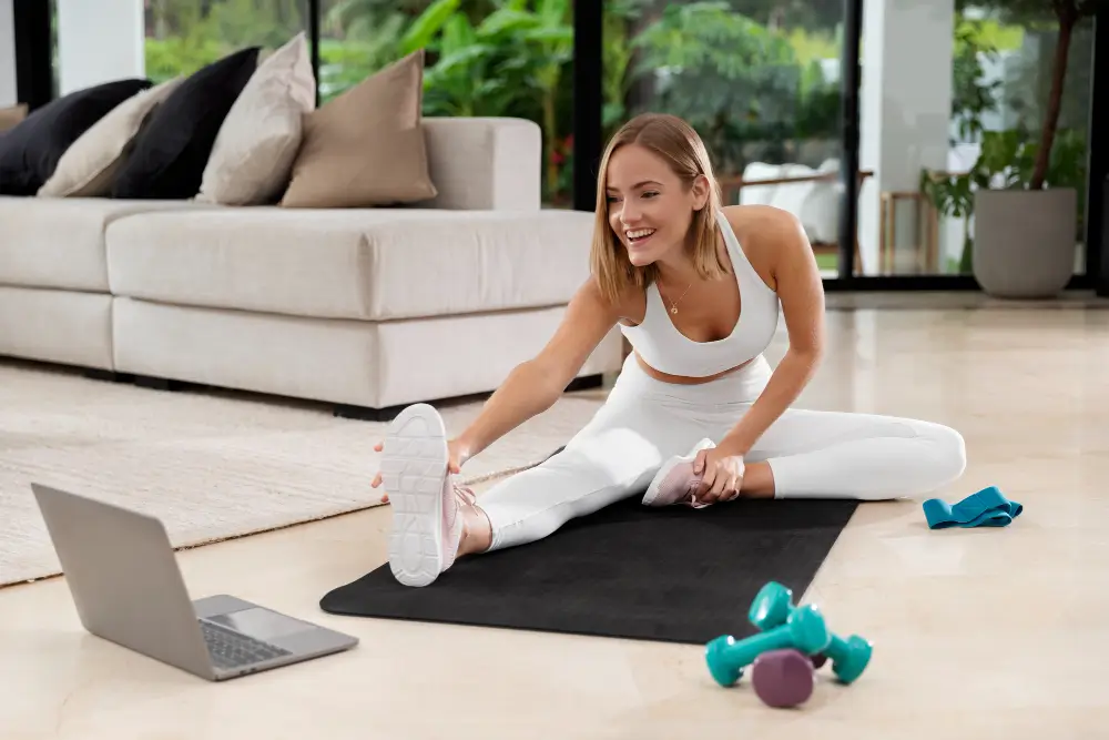 Woman stretching on yoga mat during virtual personal training session with laptop and dumbbells