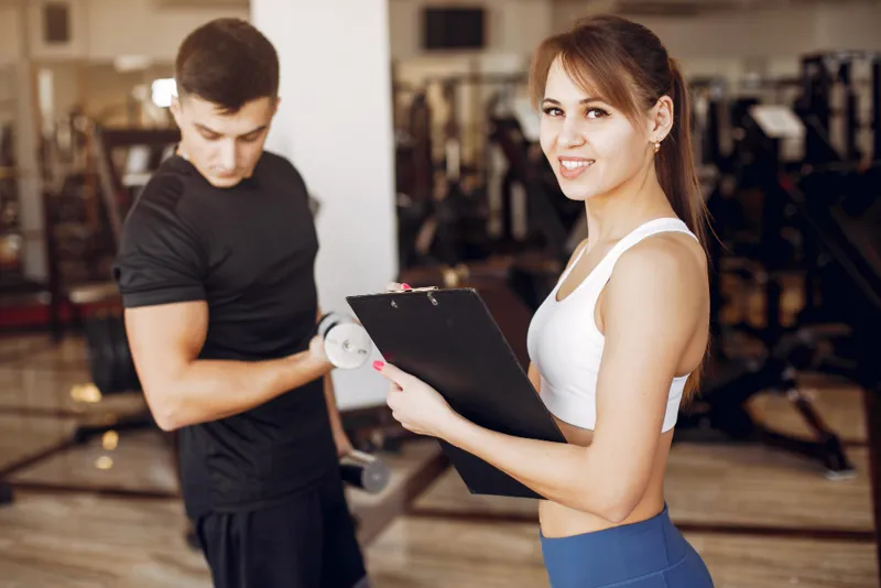 Female personal trainer holding a clipboard while smiling at the camera, as a male client lifts a dumbbell in a gym setting.
