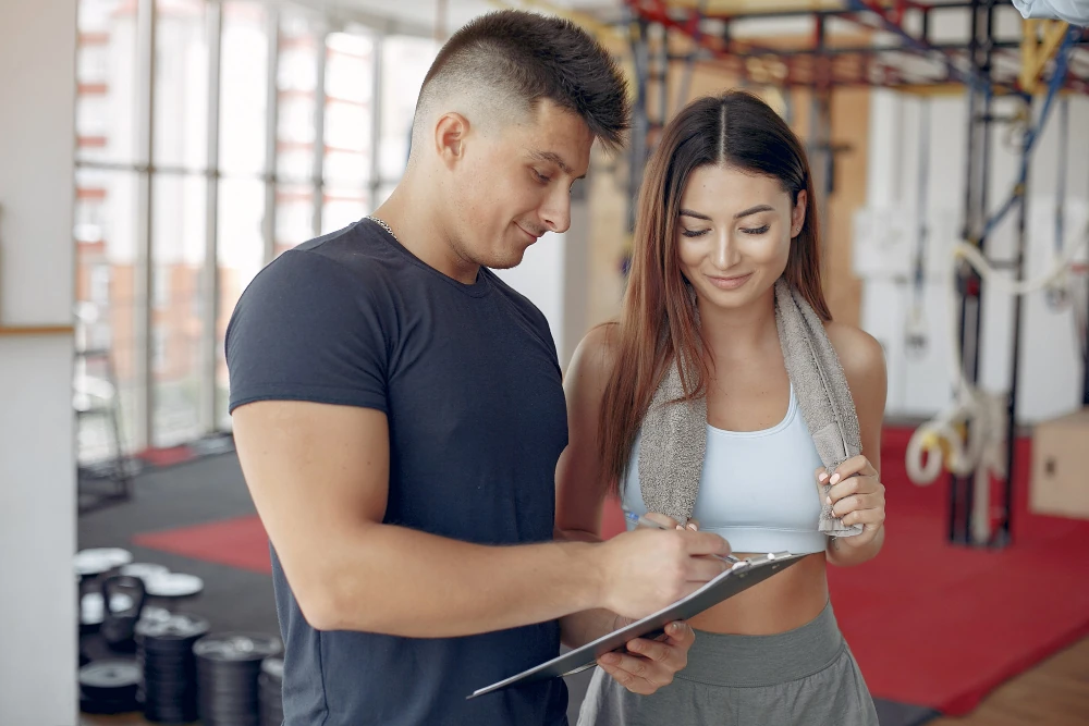A personal trainer coaching a female client in a gym, discussing a workout plan using a clipboard, representing professional fitness coaching and client management for Superset App context.