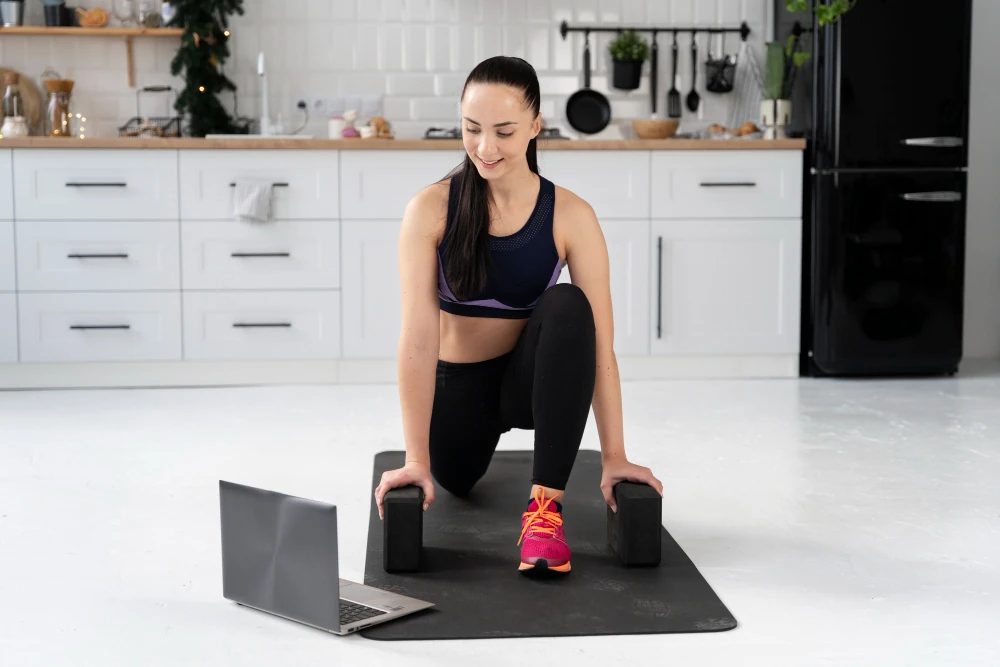 A woman exercising at home on a yoga mat with dumbbells, following an online workout on her laptop in a bright modern kitchen, representing online coaching and fitness training